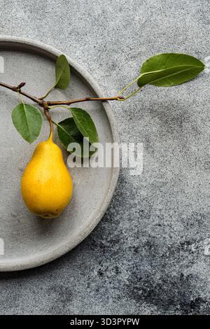 Top view of a yellow pear resting on a gray ceramic plate accompanied by a leafy branch. The setting has a minimalist style with a textured gray concr Stock Photo