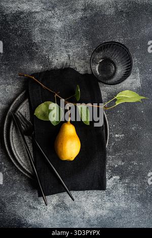 Top view of a pear with a branch on a black napkin, set on a textured gray plate. Black cutlery and a ribbed glass complement the minimalist stone con Stock Photo