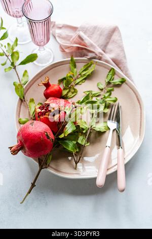 From above, a beige ceramic plate holds a pomegranate with green leaves. Pink-handled cutlery and pink glassware complement a soft pink napkin on a li Stock Photo