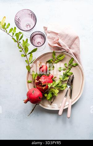Top view of a pomegranate-themed table setting featuring a beige plate with vibrant pomegranates, lush greenery, two pink glasses, and pink-handled cu Stock Photo