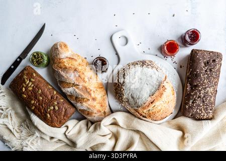 Top view of a variety of freshly baked sourdough bread loaves with seeds, accompanied by jars of colorful spreads on a textured white background and b Stock Photo