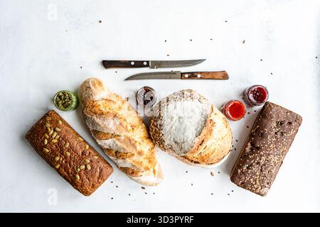 Top view of an assortment of sourdough breads including seeded and classic varieties, accompanied by colorful spreads in small jars on a light backgro Stock Photo