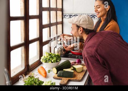 A happy couple enjoys cooking together in their bright Ecuadorian kitchen, basking in the natural light from large windows, surrounded by fresh vegeta Stock Photo