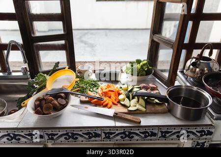 A variety of fresh vegetables including carrots, zucchini, and broccoli are neatly arranged on a cutting board beside a wooden framed open kitchen win Stock Photo
