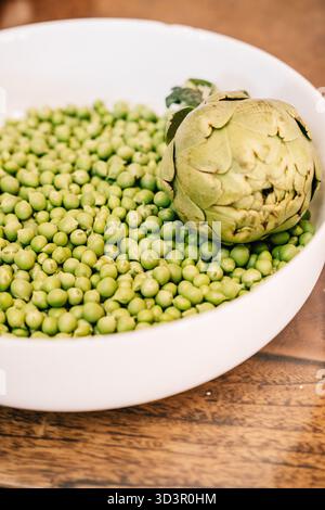 Close-up of a fresh artichoke and numerous green peas in a white ceramic bowl, showcasing vibrant green hues, perfect for culinary preparation and hea Stock Photo