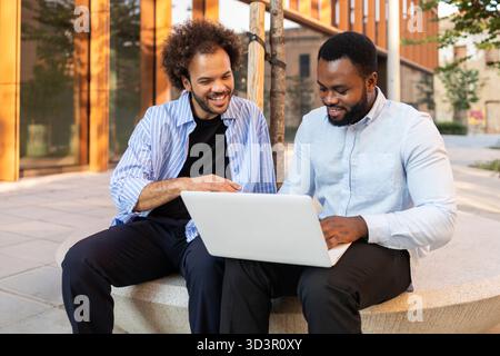 An energetic business team sits outdoors in a modern city environment, actively collaborating on a project using a laptop. They appear engaged and foc Stock Photo