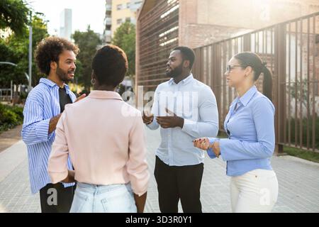 A diverse business team engages in a lively discussion outdoors in a city environment. The group stands together, sharing ideas under the sun, enhanci Stock Photo