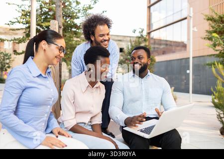 A diverse business team collaborates outdoors in an urban setting, using a laptop for their discussion. They are focused, engaged, and smiling, reflec Stock Photo
