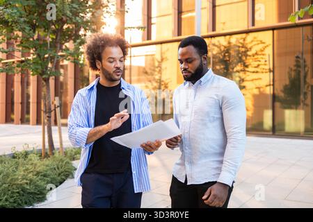 Two professionals engaging in a discussion while reviewing documents outside a modern office building in the city, highlighting teamwork and collabora Stock Photo