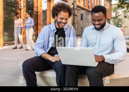 A diverse business team enjoys a productive outdoor meeting in the city. Two smiling professionals share ideas on a laptop, while others engage in con Stock Photo