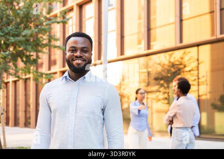 Smiling businessman stands confidently outside a modern office building, while his diverse team engages in discussion, reflecting collaboration and su Stock Photo