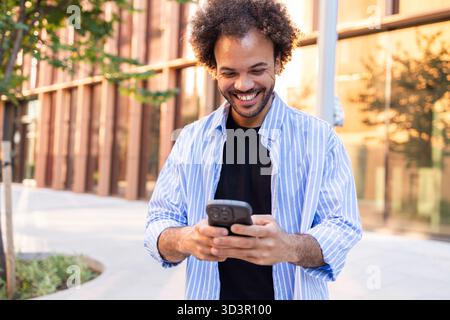 A cheerful professional stands outdoors, engaging with a smartphone in an urban setting. The scene reflects modern business dynamics, showcasing conne Stock Photo
