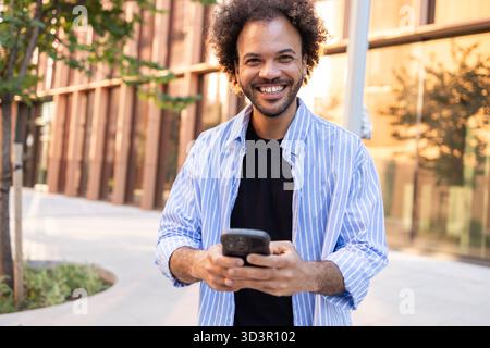 A cheerful professional stands outdoors, engaging with a smartphone in an urban setting. The scene reflects modern business dynamics, showcasing conne Stock Photo