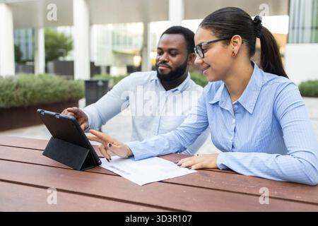 A diverse business team collaborates outdoors in an urban setting, discussing a project using a digital tablet. The city backdrop highlights modern wo Stock Photo