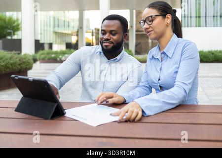 A professional business team collaborates outside in a modern city environment, engaging in strategic planning using a tablet and documents to achieve Stock Photo