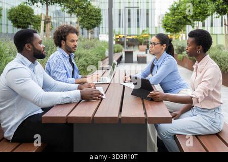 A diverse business team engages in a productive outdoor meeting at a modern urban location. They work collaboratively with laptops and tablets on a wo Stock Photo