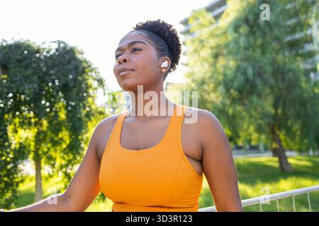 A woman runner in an orange sports top enjoys a sunny day outdoors, taking a break during her workout. The green park surroundings add a serene atmosp Stock Photo