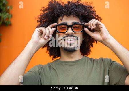 A fashionable man with curly hair smiles while adjusting his trendy sunglasses. The vibrant orange background enhances his urban and modern lifestyle Stock Photo