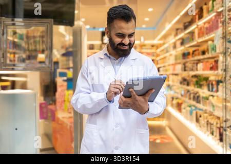 A pharmacist in a white coat uses a tablet in a modern pharmacy. Shelves stocked with medications and health products line the background, emphasizing Stock Photo