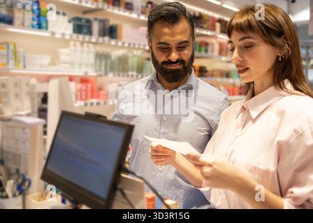 Two customers stand in a brightly lit pharmacy, discussing their purchase options. Shelves stocked with various health and beauty products are visible Stock Photo
