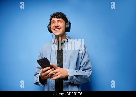 A young man wearing headphones smiles while holding a smartphone against a solid blue background. He is dressed in a blue denim shirt and appears rela Stock Photo