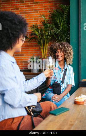 Two friends share a moment over drinks in a lively downtown setting. Surrounded by vibrant plants and warm brick walls, they enjoy a sunny day filled Stock Photo