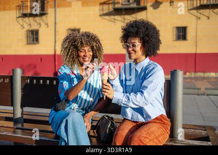 Two friends share a joyful moment while enjoying an ice cream cone in a sunny downtown area. They sit on a bench, smiling and savoring the sweet treat Stock Photo