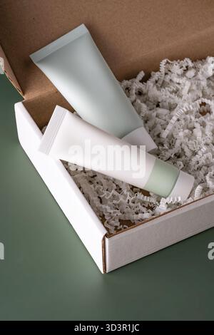 From above, this image shows two cream tubes placed in an open cardboard box filled with white packing shreds, set against a green background for an a Stock Photo