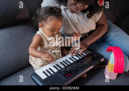 African American mother and daughter spending quality time together at home, sitting on the couch and playing a small electronic keyboard, learning an Stock Photo