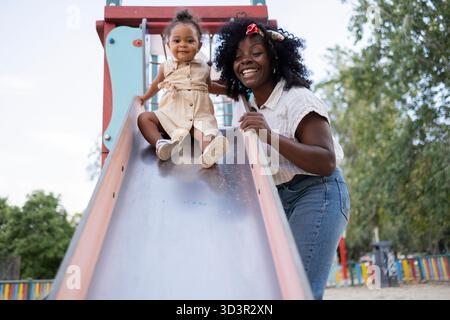 African American mother and daughter enjoying a playful day at the park. The mother smiles brightly, encouraging her daughter, who is about to slide d Stock Photo
