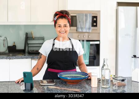 Smiling woman in an apron mixes ingredients for handmade bread in a modern kitchen. She is surrounded by tools and ingredients, reflecting a cozy home Stock Photo