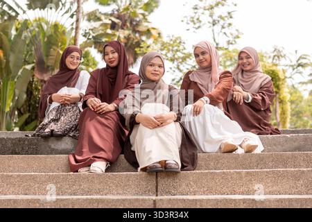 A group of young Muslim women, part of Generation Z, sit together on stone steps outdoors, enjoying friendship and wearing hijabs. The scene captures Stock Photo