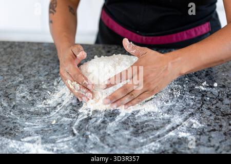A woman kneads dough on a kitchen counter, preparing homemade bread. Her hands are skillfully shaping the dough, surrounded by flour, emphasizing the Stock Photo