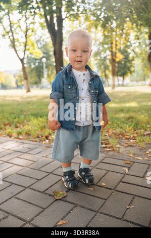 A small child stands confidently on a paved path, dressed in casual denim and light shorts, embracing the freedom and joy of nature during a sunny day Stock Photo