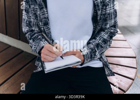 A young man sits on a curved wooden bench, jotting down notes in a notebook with a pen. Dressed casually, he appears focused and engaged in his writin Stock Photo