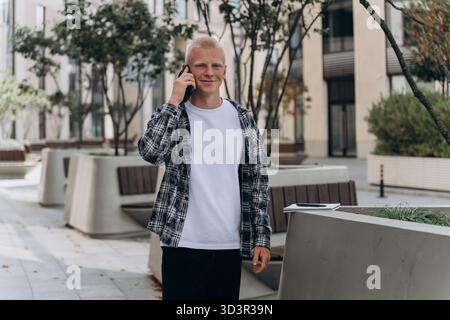 A young man with an albino appearance is standing outdoors, smiling while talking on his phone. He is wearing a plaid shirt and white t-shirt in an ur Stock Photo