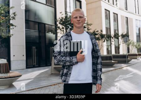 A young man with an albino appearance stands confidently outside in an urban setting, holding a notebook. He is dressed casually in a plaid shirt over Stock Photo