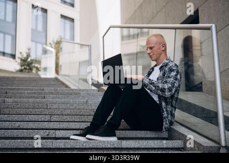 A young man with an albino appearance sits on urban steps while working on his laptop. He wears casual attire, and the scene reflects a peaceful, prod Stock Photo