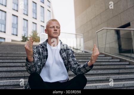 A young man with an albino appearance sits cross-legged on urban steps, meditating with a serene expression. The modern cityscape in the background ad Stock Photo