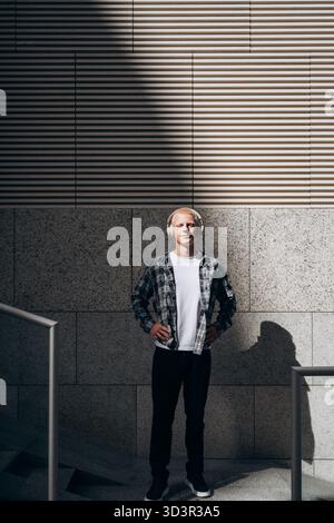 A young man with an albino appearance stands confidently in an urban setting, wearing headphones. The light casts a distinct shadow, highlighting his Stock Photo