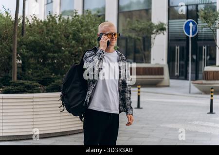 Young man with albino appearance talks on his smartphone while standing outdoors in a modern urban setting. He wears fashionable casual attire and car Stock Photo