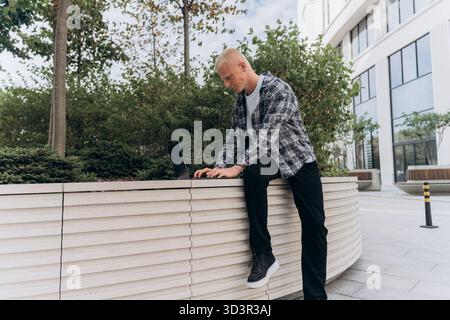 A young man with an albino appearance is focused on his laptop while leaning on a modern, urban planter in a city environment, exuding concentration a Stock Photo