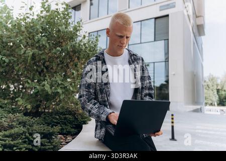 A young man with an albino appearance sits outside a modern office building, focused on working with his laptop. The setting is calm, with greenery an Stock Photo