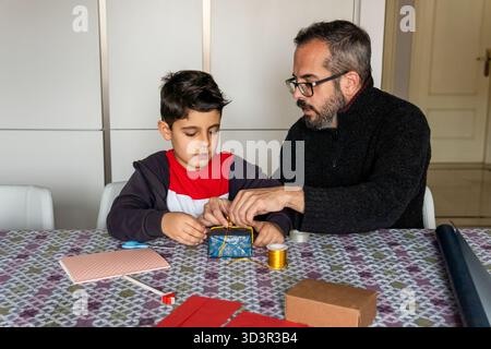 Father and son bond by wrapping a holiday gift together at home. The festive season is highlighted with a focus on family and the joy of giving, evoki Stock Photo