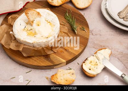 From above, the warm and creamy baked Camembert is presented on a wooden platter with crispy toasted bread slices. A sprig of rosemary adds a touch of Stock Photo