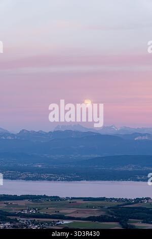 A mesmerizing full moon casts its glow over the serene Lake Geneva, with picturesque distant mountains providing a stunning backdrop against a soft, c Stock Photo