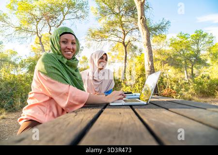 Two muslim women engage in podcasting at a tranquil park, using a laptop on a wooden table. The picturesque setting enhances their creative outdoor wo Stock Photo