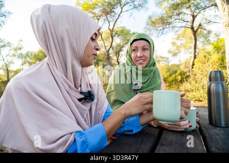 Two muslim women engage in podcasting in a tranquil park setting. They sit at a wooden table amid lush greenery, using microphones while enjoying a pe Stock Photo