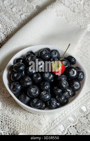 A white bowl filled with vibrant blueberries and a single strawberry on top, set on an elegant lace fabric background. Perfect for healthy food and nu Stock Photo