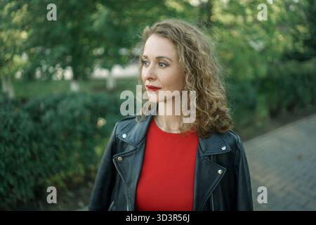 A woman wearing a stylish black jacket stands gracefully in a vibrant city park. Surrounding her are lush green trees and a serene pathway, enhancing Stock Photo